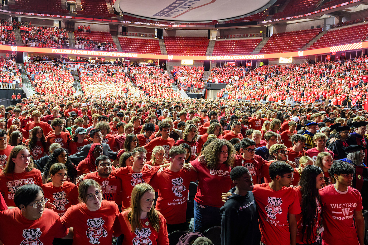 A diverse crowd of students wearing red Badger gear and with their arms around each other, sing "Varsity" at a convocation event